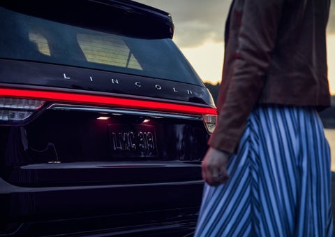 A person is shown near the rear of a 2024 Lincoln Aviator® SUV as the Lincoln Embrace illuminates the rear lights | Parks Lincoln of Tampa in Tampa FL